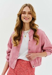Jasnoróżowa kurtka bomberka z detalami w formie dziurek, srebrnymi zatrzaskami i elastycznymi mankietami, zestawiona z białą bluzką i spódnicą w kwiatowy wzór.