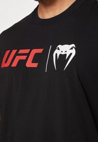 Černé bavlněné tričko se výrazným červeným logem "UFC" a bílým emblémem hada. Standardní kulatý krk a měkká textura.
