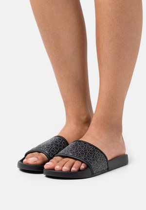 Pieds portant des sandales noires à enfiler avec une bride à motif affichant le logo "CK" répétitif sur un fond blanc.