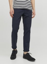 Navyblaue Baumwollchinos mit schlankem Schnitt und aufgekrempelten Bündchen, kombiniert mit schwarzen Wildlederschuhen und hellen Socken. Gestreiftes Hemd teilweise sichtbar.