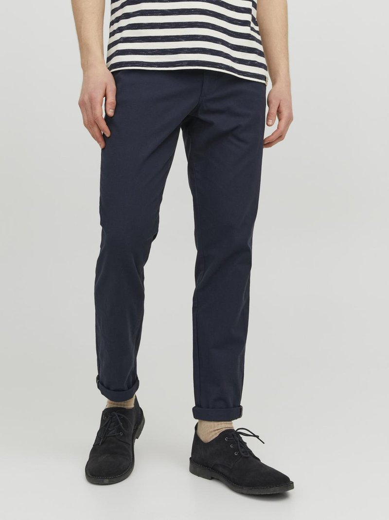Navyblaue Baumwollchinos mit schlankem Schnitt und aufgekrempelten Bündchen, kombiniert mit schwarzen Wildlederschuhen und hellen Socken. Gestreiftes Hemd teilweise sichtbar.