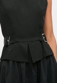 Μαύρο αμάνικο φόρεμα με ζώνη στο ύψος της μέσης, μεταλλικές αγκράφες και υφασμάτινη υφή. Πλισέ στοιχείο φούστας κάτω από τη μέση.