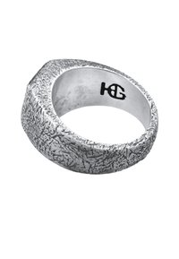 Bague de couleur argentée avec une surface texturée et rugueuse, de forme arrondie. À l'intérieur, elle présente un logo noir gravé.