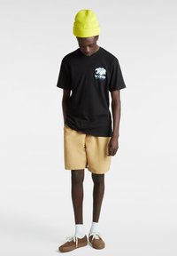 Camiseta negra con un gráfico de palmera, pantalones cortos beige, calcetines blancos y zapatos marrones. Gorro amarillo brillante. Conjunto casual con colores contrastantes.