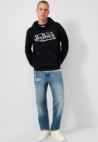 Zwarte hoodie met witte "Von Dutch" print, denim jeans met logo patch en witte sneakers. Casual outfit met een relaxte pasvorm en minimale details.