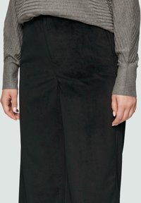 Czarne spodnie typu wide-leg wykonane z miękkiego, teksturowanego materiały, zestawione z szarym, prążkowanym topem z długim rękawem. Widoczna ręka z jasnym lakierem do paznokci.