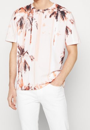 Mężczyzna ubrany w białe spodnie i białą koszulę z krótkim rękawem, wzór z brzoskwiniowymi i brązowymi palmami, jedna ręka w kieszeni.