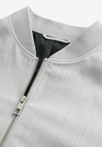 Jachetă gri deschis, cu închidere prin fermoar și guler ribat, dintr-un material texturat și căptușită cu plasă în interior. Fermor argintiu.