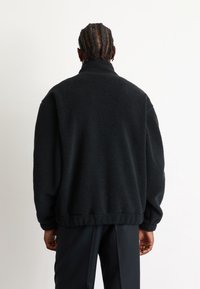 Chaqueta de forro polar en negro, cuello alto, corte holgado, tejido texturizado, puños acanalados. Combinada con pantalones negros, vista desde la parte posterior.