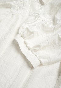 Blusa bianca in tessuto di lino testurizzato con intricato ricamo in pizzo lungo lo scollo e maniche a sbuffo con polsini elasticizzati.