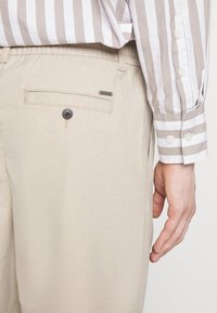 Beige chinos s zadními náprsními kapsami s knoflíkem, spárované s lehce pruhovanou košilí s dlouhým rukávem a manžetami na knoflíky.