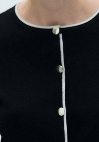 Černý pletený kardigan s kulatým výstřihem, který má pět stříbrných knoflíků a bílý lem podél okraje a knoflíkové légy.