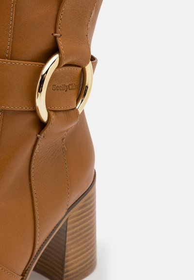 Brun læder ankelstøvle med en bred rem og en guldfarvet, rund spænde. Har en kraftig, struktureret træhæl.