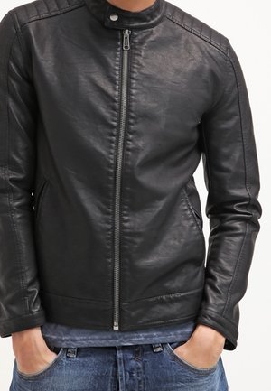 Mężczyzna ubrany w dopasowaną czarną skórzaną kurtkę z zamkiem błyskawicznym z przodu i zapinanym na zatrzask kołnierzem, ręce w kieszeniach niebieskich dżinsów.