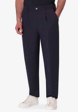 Uomo con pantaloni dritti blu scuro con tasche laterali e sneakers bianche con lacci, in piedi su uno sfondo plain.