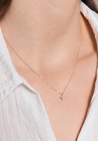 Złoty łańcuszek z małym zawieszeniem w kształcie gwiazdki z przezroczystym kamieniem w środku, noszony na nagiej szyi, na tle jasnej koszuli.