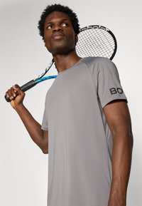T-shirt de sport gris à manches courtes, en tissu texturé, avec un design raglan. Détail du logo noir sur la manche. Raquette de tennis tenue sur l'épaule.