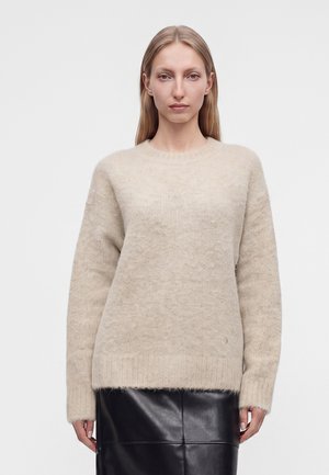 Femme aux cheveux blonds et raides portant un pull beige duveteux et une jupe en cuir noire, debout devant un fond clair uni.
