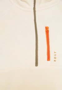 Polarowy pulower w kolorze off-white z pół-zamkiem. Posiada kontrastowy pomarańczowy suwak oraz delikatne, złote logo z boku.