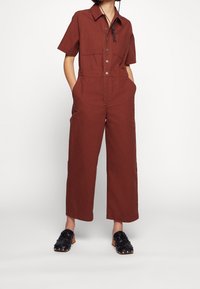 Jumpsuit w kolorze rdzy z krótkimi rękawami, dwoma kieszeniami na piersiach, zapięciem na guziki z przodu i szerokimi nogawkami. Model połączył go z czarnymi plecionymi mule.