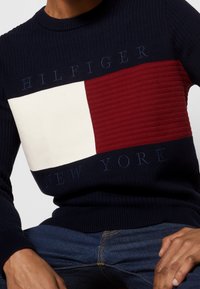 Pull en maille bleu marine avec une texture côtelée, présentant un large panneau horizontal rouge et blanc, affichant "HILFIGER NEW YORK" en bleu marine.