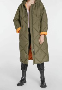 Olivgrön vadderad lång kappa med huva, med orange foder, diamantformigt quiltmönster och dragkedja framtill. Svarta ankelstövlar.