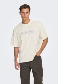 Kremowy oversize t-shirt z okrągłym dekoltem i krótkimi r�ękawami, na przodzie napis „Très Bien” w szarym stylu kursywy.