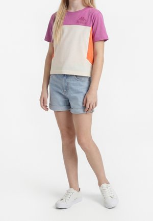 Barn iført en t-shirt med farveblokke i lilla, beige og orange, lyse denim rullede shorts og hvide sneakers, der står mod en hvid baggrund.