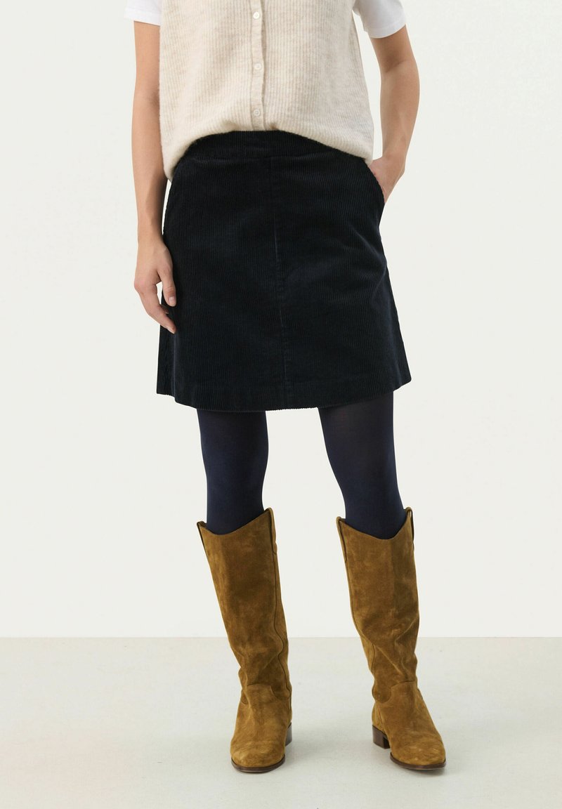 Dark corduroy skirt with a straight cut, paired with a cream knit top and knee-high brown suede boots. Tights in a dark color complement the outfit.