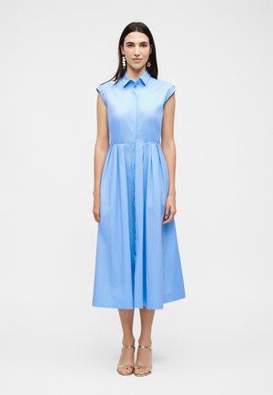Kobieta stojąca, ubrana w bezrękawnikową jasnoniebieską sukienkę midi z kołnierzykiem i przednimi guzikami, zestawioną z beżowymi sandałami na wysokim obcasie oraz kolczykami.
