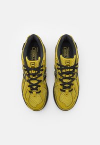 Žluté sportovní boty s černými detaily, texturovaným síťovaným svrškem, klasickým šněrováním a gumovou podrážkou. Číslo modelu "1906" je viditelné na jazyku.