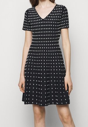 Sort kortærmet V-halskjole med hvide polkaprikker, tætsiddende talje og plisseret nederdel, båret af en stående person.