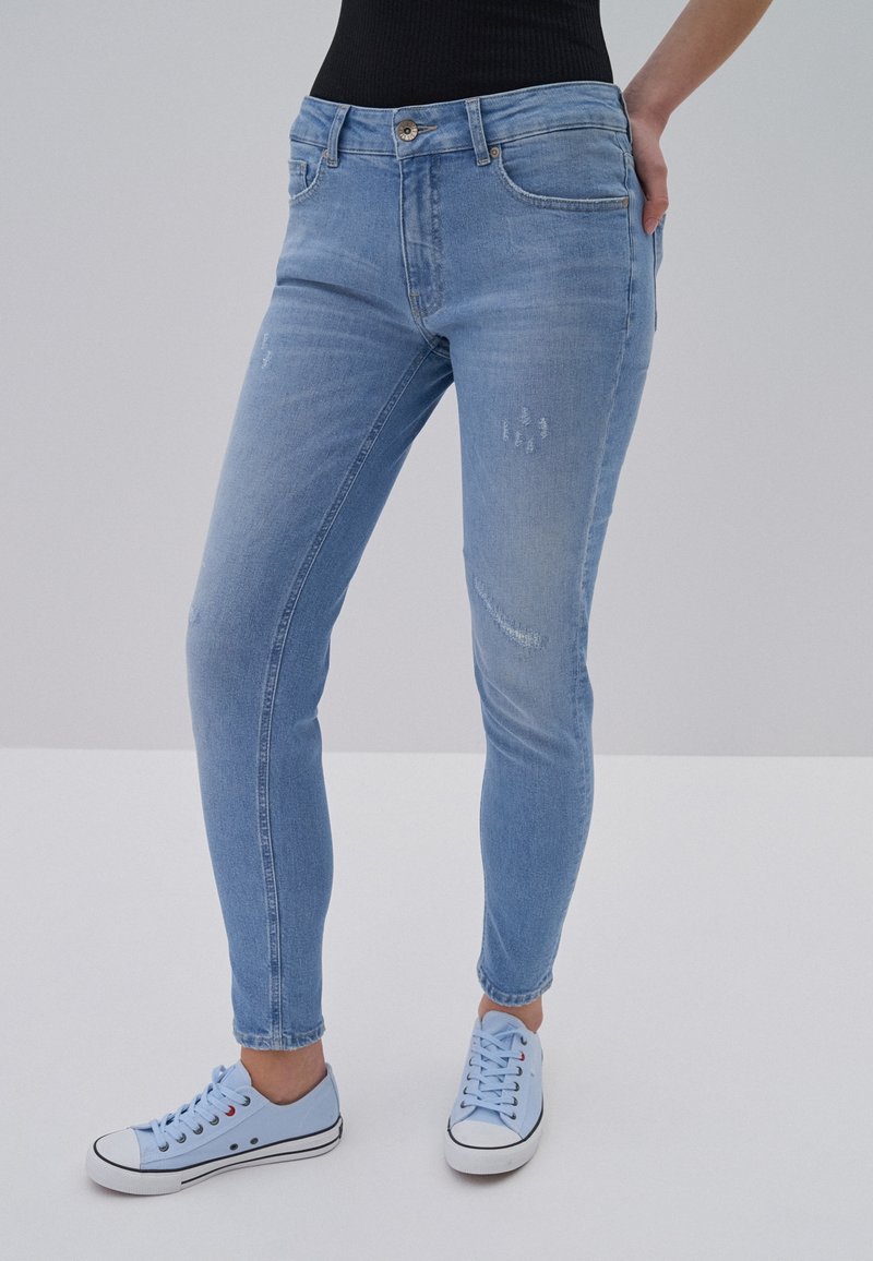 Femme portant un jean skinny bleu clair avec légère usure et des baskets en toile bleu clair à lacets, main reposant sur la hanche.