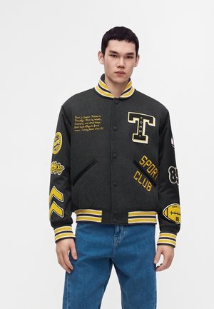 Jeune homme portant une veste universitaire noire et jaune avec des écussons et du texte, associée à un jean bleu, debout devant un fond blanc.
