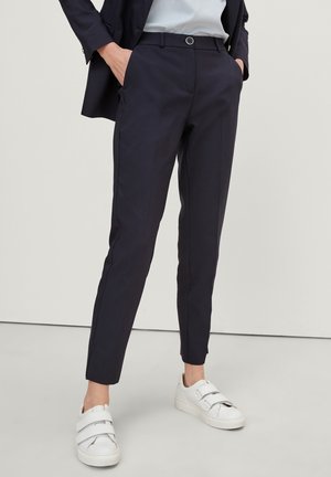 Persoon draagt slim fit donkerblauwe broek, zwarte blazer, lichtblauw topje en witte klittenbandsneakers, staand met handen in de zakken.