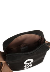 Borsa a spalla in tessuto nero con interno marrone; presenta una chiusura con zip e un grande logo bianco sul davanti; tracolla regolabile visibile.