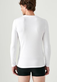 Witte langemouwen shirt te zien vanuit de achterkant, gemaakt van gladde stof, met een klassieke ronde halslijn en een aansluitend ontwerp, gecombineerd met zwarte shorts.