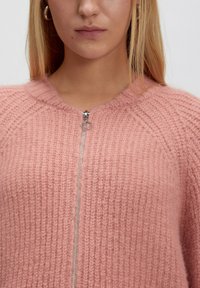 Hellrosa, gestrickter Pullover mit strukturiertem, geripptem Muster. Verfügt über einen Frontreißverschluss mit einem runden Ziehgriff und einem runden Halsausschnitt.
