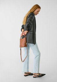 Sac en cuir marron avec un design courbé, une bandoulière ajustable et une finition élégante. Associé à une veste noire et un jean bleu clair.