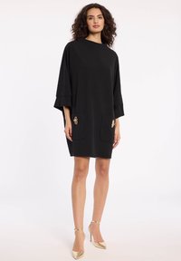 Schwarzes Kleid aus glattem Stoff, mit Fledermausärmeln und zwei vorderen Taschen mit goldenen Akzenten. Oberhalb des Knies geschnitten.