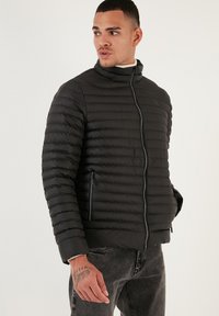 Giacca a vento nera imbottita con design a righe orizzontali, colletto alto, chiusura frontale con zip e tasche laterali. Tessuto liscio, materiale leggero.