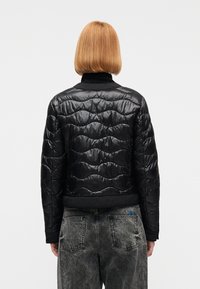 Άτομο με κοντά ίσια κόκκινα μαλλιά, φορώντας μαύρο καπιτονέ μπουφάν και ξεθωριασμένο μαύρο τζιν, από πίσω, σε απλό φόντο.