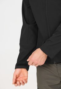 Gros plan d'une personne ajustant la manche d'une veste noire avec une sangle en Velcro, portant un pantalon olive, sur un fond uni clair.