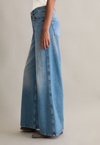 Jeans en denim bleu à jambe large avec une taille mi-haute, présentant un devant lisse, des poches latérales et une coupe droite et ample des hanches à l'ourlet.