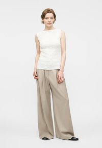 Γυναίκα που φοράει λευκό αμάνικο τοπ με υφή, φαρδιά μπεζ παντελόνια και μαύρα παπούτσια, στέκεται μπροστά σε απλό ανοιχτό φόντο.