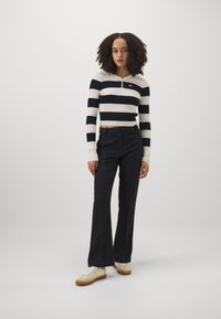 Rästripäevitav cropped-sviiter kreemi ja tumesinise värviga, ribakujuline tekstuur; koos mustade kohandatud pükste ja valgete sneakeritega, millel on kollased aktsendid.