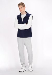 Navy modrá zipsová vesta cez biele tričko s dlhým rukávom, doplnená sivými teplákmi a bielymi teniskami. Jednoduchý dizajn a ľahká textúra látky.