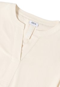 Collo in V di una camicia di colore crema con etichetta bianca visibile che reca la scritta "CECIL" su sfondo bianco.