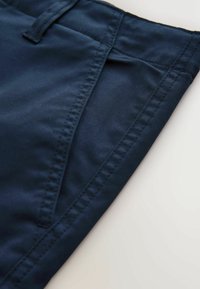 Prim-plan cu buzunarul unei perechi de pantaloni bleumarin, cu cusături vizibile și buclă pentru curea, pe un fundal deschis.