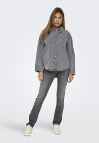 Camicia di denim grigia oversize con bottoni, abbinata a jeans grigi slim. Il modello indossa sneakers chiare, mostrando un outfit casual e monochromatico.